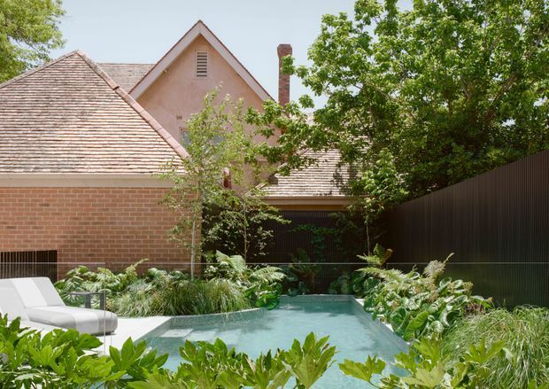 A landscaped courtyard garden with a small swimming pool in the foreground, surrounded by lush planting, ornamental grasses, and a white outdoor lounge chair. Behind the garden is a traditional brick and rendered residential building with steep pitched, shingled roofs, gabled forms, a chimney, and a dark vertical fence. The scene is bright and leafy with a tranquil, private atmosphere.