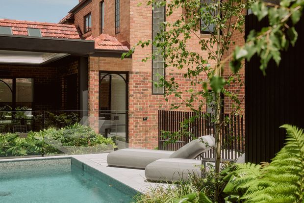 A landscaped courtyard pool beside a multi-storey red brick residence. The scene features a covered outdoor area with dark framing, a tiled terracotta roof, arched glazing and doorway details, tall narrow windows, timber screening, and two light-coloured sun loungers along the pool edge. Lush planting surrounds the turquoise swimming pool, creating a private, leafy outdoor setting.