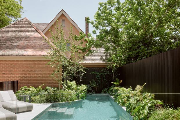 A landscaped residential courtyard with a narrow rectangular swimming pool, bordered by lush planting and a dark vertical metal privacy fence. The pool sits beside a red brick outbuilding with a pitched terracotta-tiled roof and a taller gabled brick structure beyond. A pale outdoor lounge chair is partially visible in the foreground. The scene has a refined, leafy inner-city garden atmosphere with strong architectural detailing and abundant greenery.