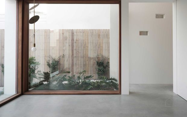 Minimalist interior with a polished concrete floor, white walls, and a large dark timber-framed corner window overlooking a planted courtyard with a timber privacy fence. A sculptural hanging pendant is visible near the glazing, while a simple hallway or adjoining room opens to the right.