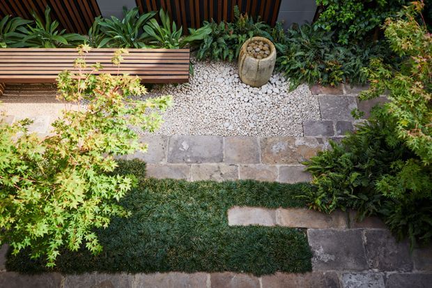 Top-down view of a compact landscaped courtyard or garden, featuring irregular stone paving, a pale gravel bed, dense layered planting, a slatted timber bench, and a stone planter or water feature. The composition has a natural, textural contemporary garden design.