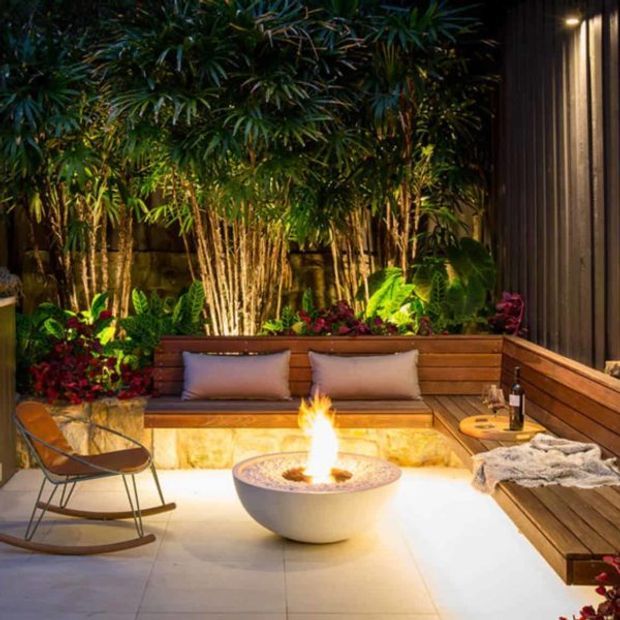 A warmly lit, intimate outdoor courtyard lounge at night, featuring built-in timber bench seating with cushions, a round fire bowl, a rocking chair, pale tiled paving, and lush tropical planting enclosed by timber-clad walls.
