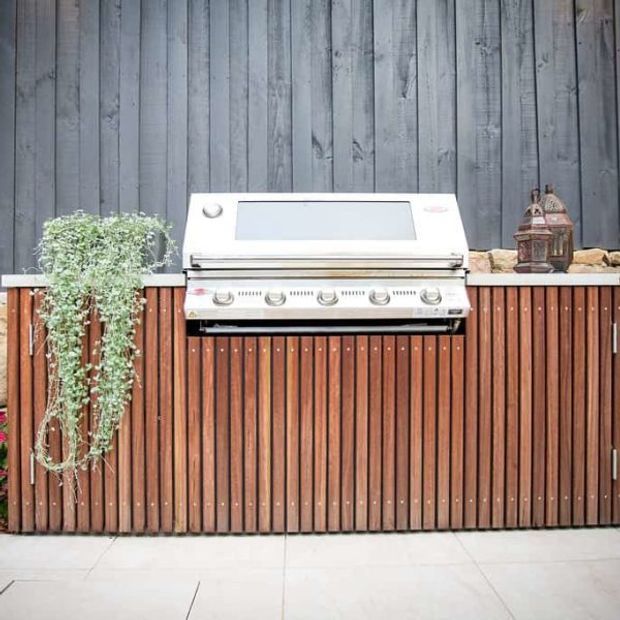 A built-in outdoor barbecue grill set into a timber-clad outdoor kitchen unit. The cabinetry has vertical warm-toned wood slats, a light stone or solid-surface benchtop, and a dark grey vertically boarded wall behind it. A trailing plant and decorative lantern sit on the bench.