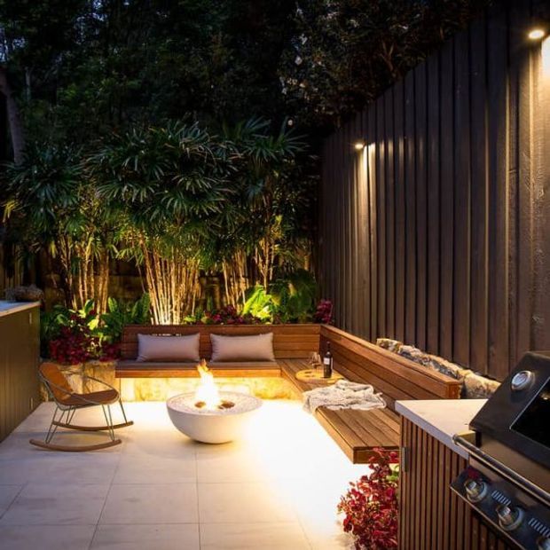 A warmly lit, intimate outdoor entertaining area with large-format tiled paving, built-in timber bench seating, a central fire bowl, lush tropical planting, and dark vertical-clad walls. A built-in outdoor kitchen or barbecue is visible at the right, with integrated wall and garden lighting creating a cozy evening atmosphere.