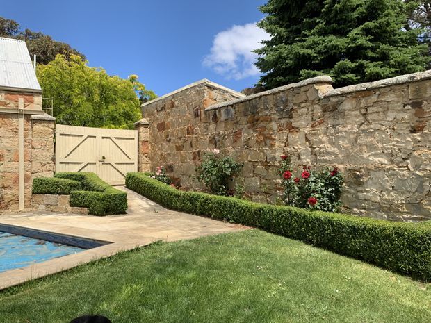 A landscaped historic courtyard garden with a stone perimeter wall, clipped box hedges, rose bushes, lawn, paved poolside area, and a pale timber double gate. Weathered masonry outbuildings and a corrugated metal roof are visible behind the wall under a clear blue sky.