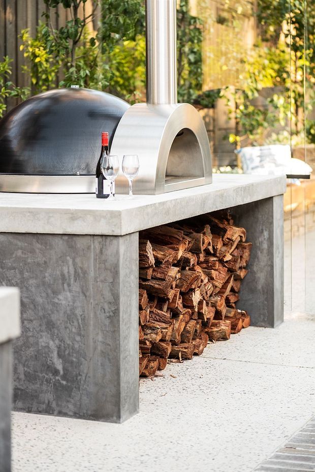 A built-in outdoor kitchen or entertaining station featuring a concrete island with an integrated wood-fired pizza oven, chimney, dark dome-shaped oven, and an open wood-storage niche beneath. The scene is set in a leafy, glazed outdoor area with terrazzo-like flooring and a concrete finish.