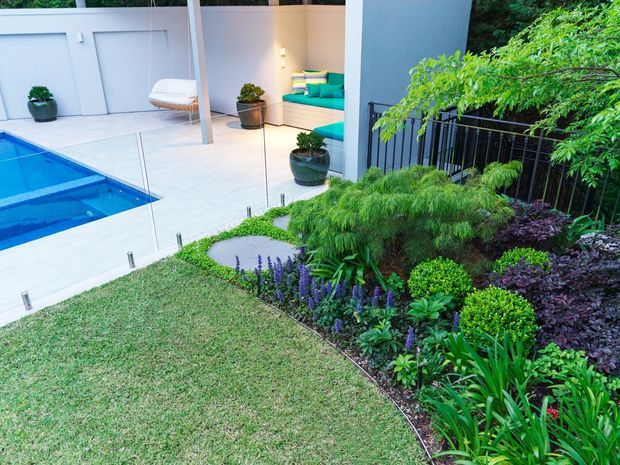 A landscaped courtyard and pool area viewed from a lawn, featuring a bright blue swimming pool, pale tiled terrace, minimalist white outdoor walls, a hanging chair, built-in turquoise seating, and a lush layered garden with ornamental grasses and flowering plants beside a black metal railing.