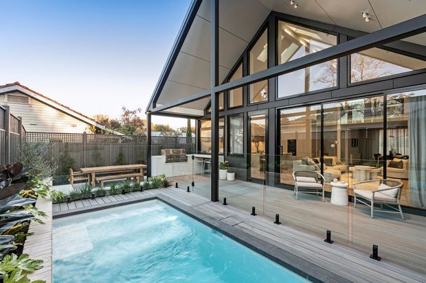Contemporary residential courtyard viewed from beside a swimming pool, featuring a timber deck, frameless glass pool balustrade, landscaped planting, and a dramatic double-height glazed pavilion. The home has a steeply pitched roof, black structural framing, extensive floor-to-ceiling glazing, and an open-plan living area visible inside.