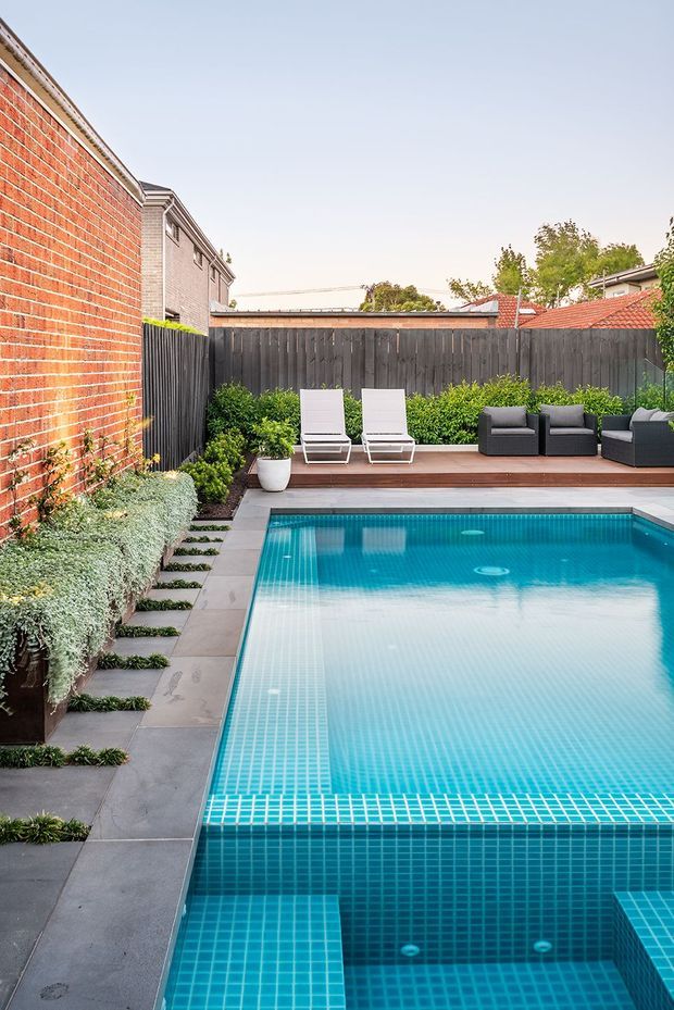A compact urban courtyard pool area with a rectangular tiled swimming pool, grey stone paving, timber decking, white sun loungers, dark outdoor sofas, raised garden beds, and dense screening greenery. Exposed red brick and neighbouring residential buildings frame the enclosed outdoor space.