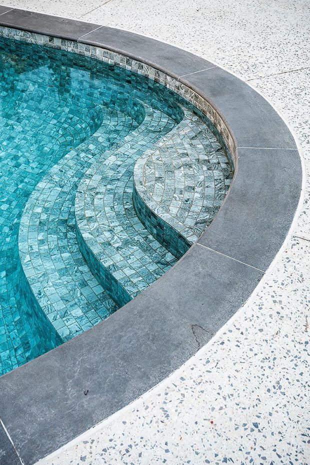 Close-up view of a curved swimming pool with turquoise mosaic tile lining, integrated curved steps or seating, and a dark grey concrete coping bordered by a light terrazzo-style paved surface.