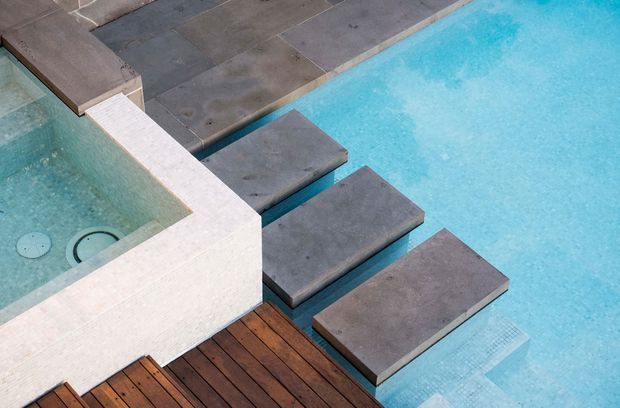 Close-up architectural view of a contemporary pool area, featuring bright turquoise water, dark grey rectangular stepping slabs extending across the pool, a pale stone-clad raised spa or pool edge, and a timber decking platform.