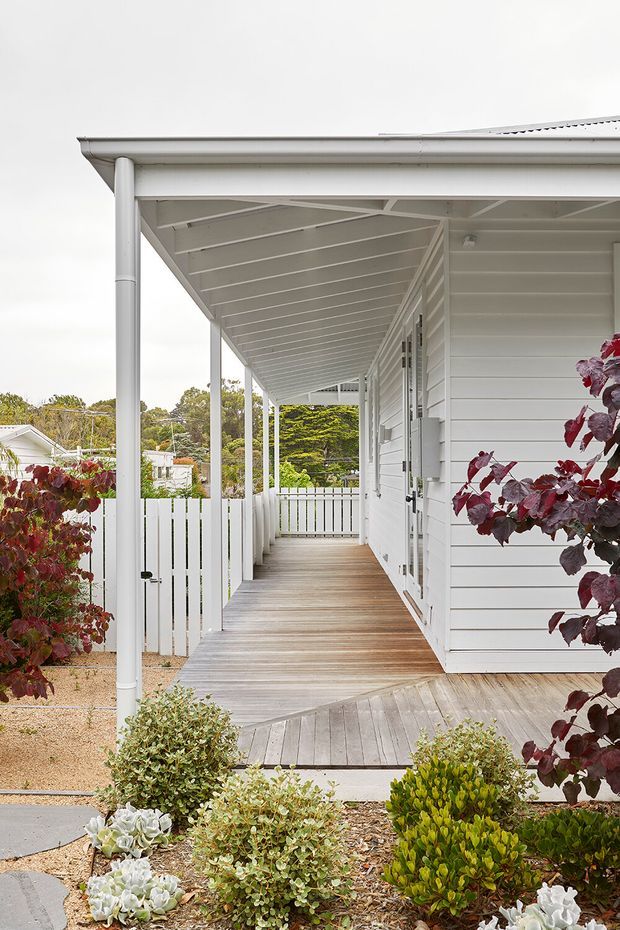 Exterior view of a white weatherboard house with a long covered verandah, timber decking, slim posts, white balustrade fencing and a pitched roof edge. A landscaped front garden with gravel, shrubs and small trees surrounds the verandah, with neighbouring homes visible in the background.