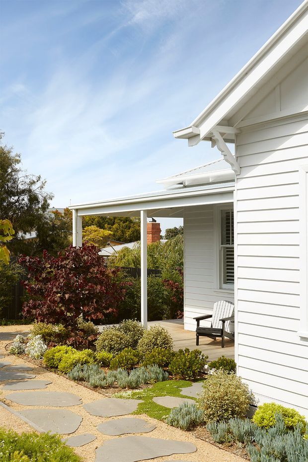Exterior view of a white weatherboard house with a pitched gable roof, covered porch, and timber decking. A landscaped front garden features low drought-tolerant plantings, gravel, and irregular stepping stones leading toward the house. The bright scene also shows a white-framed window, decorative gable detailing, and a sheltered outdoor sitting area.