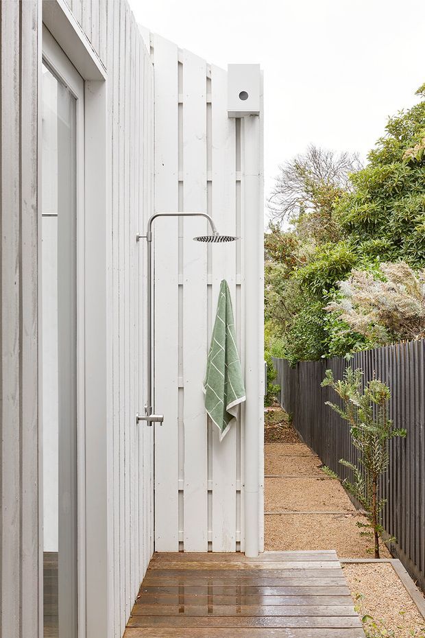 A narrow outdoor shower beside a white vertically clad exterior wall, with a simple metal shower fixture, green towel, timber decking, and a landscaped gravel side path enclosed by a dark fence. The scene has a clean, minimalist coastal-residential feel.