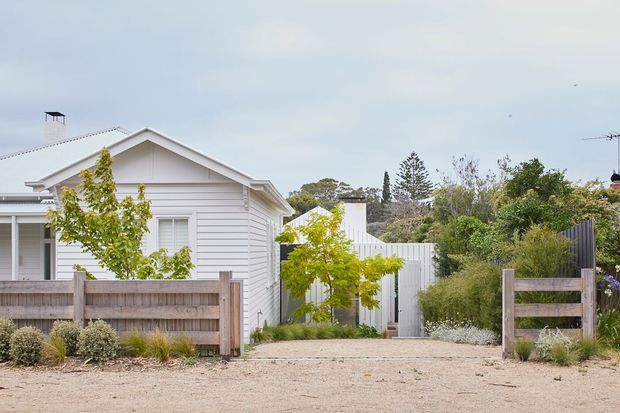 Exterior view of a light-painted horizontal weatherboard house with a prominent gabled roof, white trim, timber fencing, and a landscaped sandy front path or driveway. The residential facade is surrounded by shrubs and small trees, creating a calm coastal or suburban character.