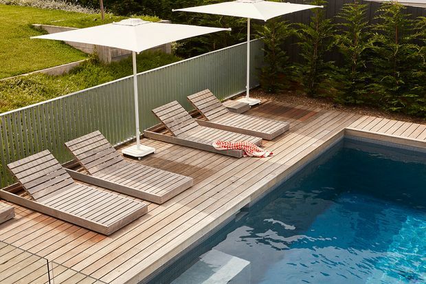 An elevated view of a private outdoor pool area with a timber deck, several slatted timber sun loungers, and large white cantilever-style umbrellas. A light-coloured privacy fence and landscaped greenery surround the deck, creating a relaxed residential outdoor setting.