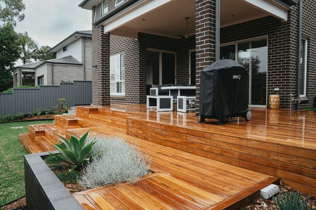 Exterior view of a contemporary brick home showing a covered rear patio and broad timber deck with built-in steps, outdoor dining furniture, and a covered barbecue. The deck connects to landscaped garden beds and a fenced yard.