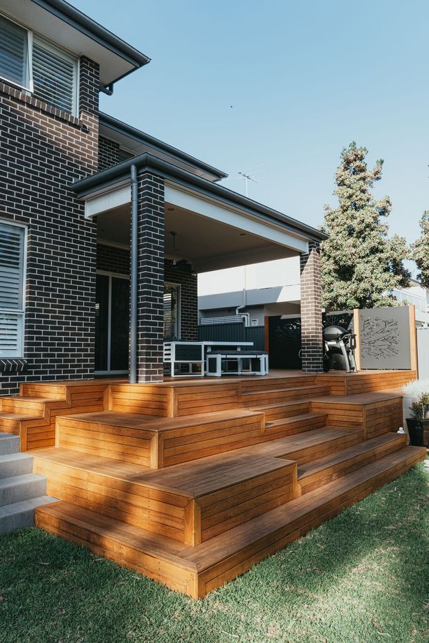 Exterior view of a contemporary brick home with a large multi-level timber deck and broad timber steps leading to a covered outdoor entertaining area. The facade features dark brickwork, white-framed louvred windows, a covered veranda supported by brick columns, metal downpipes, outdoor furniture, and a decorative privacy screen. Lawn and mature planting surround the deck.