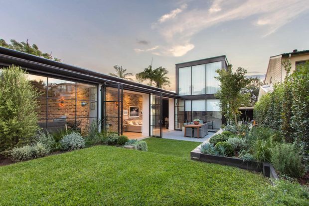 Contemporary residential courtyard viewed from the landscaped garden, featuring a low-slung dark metal roof, extensive floor-to-ceiling glazing, exposed warm brick walls, and a two-storey glazed volume. The courtyard includes a lawn, raised garden beds, mature planting, and an outdoor dining area visible through the glazing.