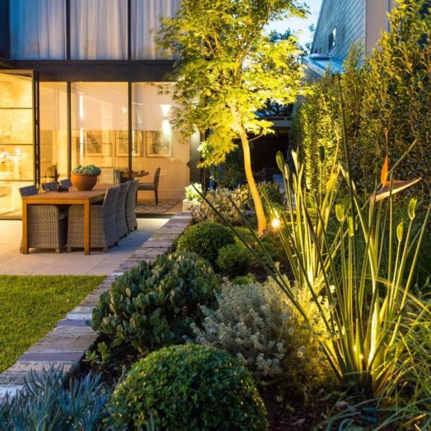 A landscaped courtyard beside a contemporary home, featuring a paved path, layered planting, a small lawn, and an outdoor dining setting beneath full-height glazed doors. The building has dark metal-framed glazing, a vertically screened upper level, and light horizontal exterior cladding. Warm evening lighting highlights the garden and patio.