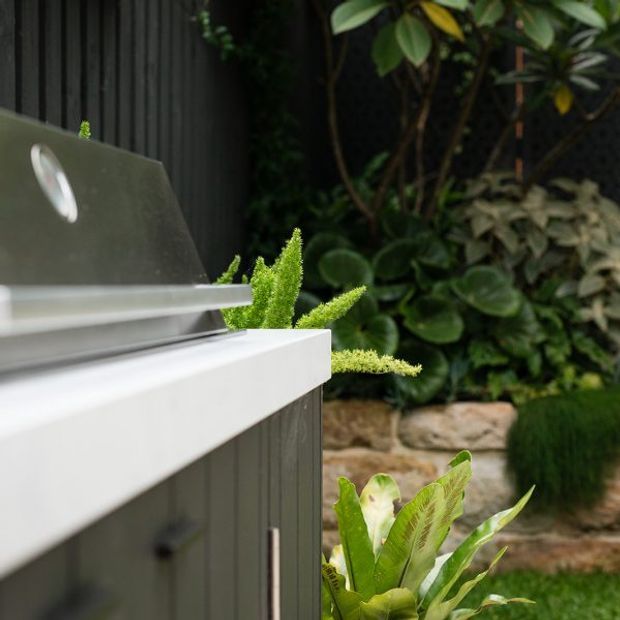 Close-up exterior view of a sleek built-in outdoor kitchen barbecue set against a lush landscaped garden. The barbecue has a dark metal lid, white benchtop, and dark vertical cladding, with layered planting and a low stone retaining wall behind it.