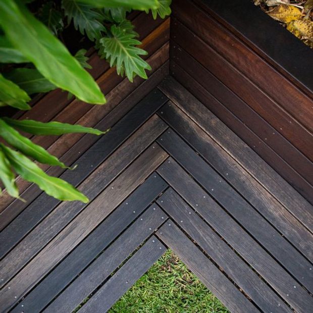 Close-up view of a dark stained timber deck corner with diagonal herringbone-style boards, matching timber wall cladding, and lush green plants along the edge. The image focuses on the outdoor decking finish and detailed joinery.