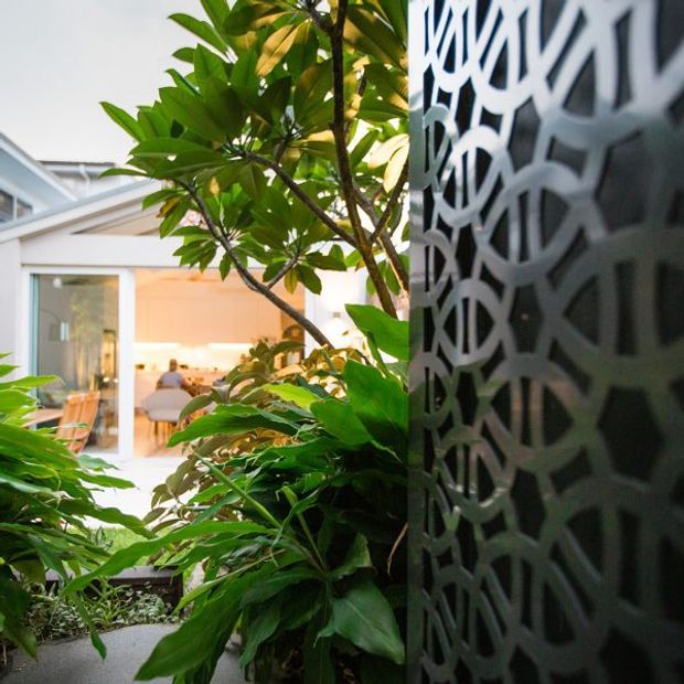 A close-up architectural view of a lush planted courtyard or entry garden, with dense tropical foliage in the foreground and a dark decorative perforated screen forming a strong vertical element on the right. Beyond the planting, a bright glazed interior space with pale walls and timber-toned finishes is visible.