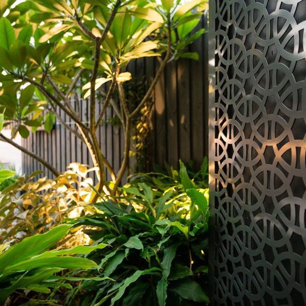 A lush landscaped courtyard or garden corner with dense tropical foliage in front of a dark vertical-slat fence. A tall dark metal decorative screen with an interlocking circular cut-out pattern forms a prominent architectural feature on the right, with warm light glowing behind it.
