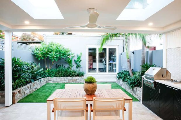 Bright covered outdoor entertaining area with a dining table opening onto a landscaped courtyard. The space features a built-in outdoor kitchen with barbecue, a white ceiling with skylights and ceiling fan, pale tiled flooring, rendered walls, raised stone garden beds, tropical planting and artificial turf.