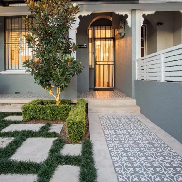 Front entry of a renovated terrace-style home, featuring a pale grey rendered facade, ornate arched porch openings, black metal security doors and window grilles, patterned tiled entry path, steps, and a neatly landscaped garden with clipped hedges and groundcover.