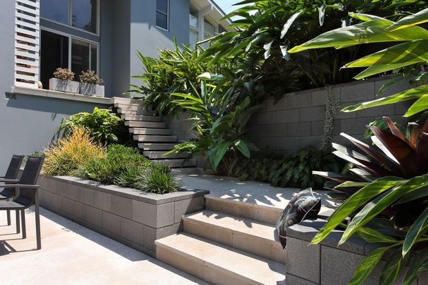 A modern landscaped courtyard or terrace beside a contemporary multi-level residence. Broad pale concrete steps connect raised garden beds, with dark textured block retaining walls, dense tropical planting, and a simple outdoor seating area. The house has grey rendered walls, large windows, and a white vertical privacy screen.