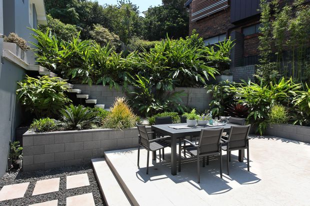 A landscaped urban courtyard with a pale paved dining terrace, outdoor table and chairs, and raised grey masonry garden beds filled with lush tropical planting. The enclosed space is bordered by modern residential facades and stepped levels, creating a private outdoor entertaining area.