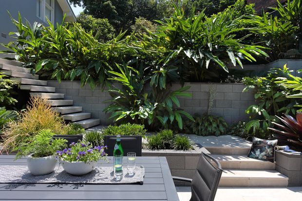 A lush, landscaped courtyard or terrace with a paved outdoor dining area, potted plants, dense tropical-style planting, and raised concrete block garden beds. Broad steps connect the dining terrace to an upper garden level, with a low retaining wall and integrated seating creating a layered outdoor space.