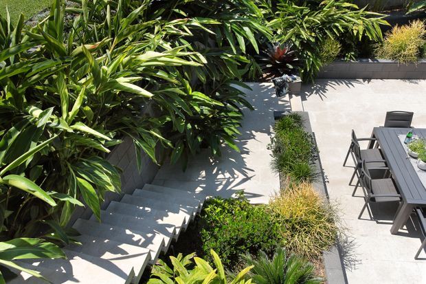 Elevated view of a landscaped outdoor courtyard or terrace with pale concrete paving, broad steps, dense tropical planting, raised garden beds, and a dark outdoor dining table with chairs.