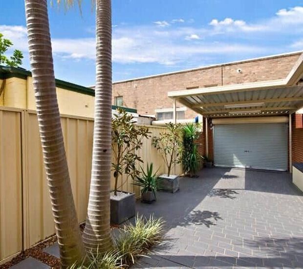 A sunny paved driveway and courtyard-style approach leading to a covered garage with a roller door. The space is enclosed by pale rendered and metal fencing, with brick building walls and tropical landscaping in raised planters.