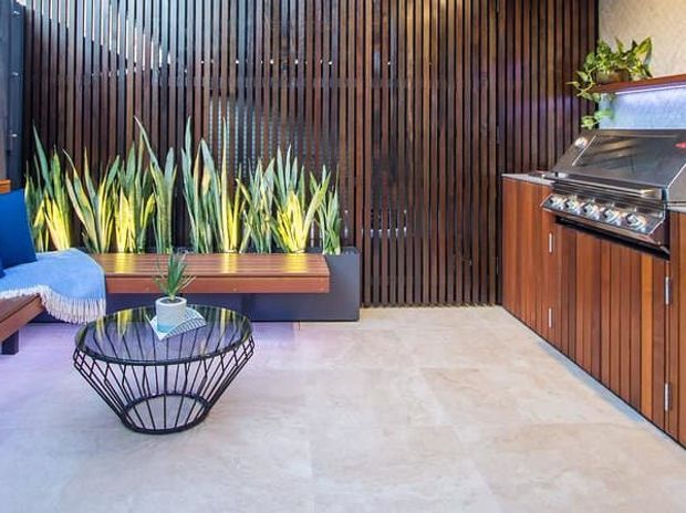 A refined outdoor entertaining area with a built-in barbecue kitchen, dark timber slatted privacy screen, integrated planter seating, and a small lounge arrangement around a sculptural black wire coffee table. Pale stone paving contrasts with the rich timber cabinetry and lush green planting.