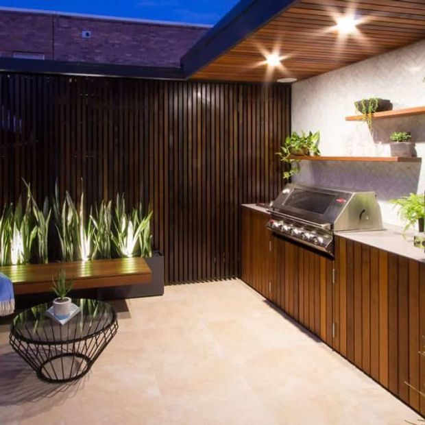 A covered outdoor entertaining area with a built-in stainless-steel barbecue, timber cabinetry, open shelving, vertical timber screening, and integrated planter seating. Warm wood tones, pale flooring, and abundant greenery create a refined, inviting outdoor kitchen and lounge setting illuminated by recessed ceiling lights.