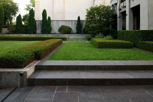 Formal landscaped courtyard or garden beside a modern building, with clipped rectangular hedges, conical evergreen topiary, lawn, concrete retaining walls, and broad dark stone steps leading through the garden. The building facade has pale rendered walls and dark vertical recesses with sculptural exterior elements.