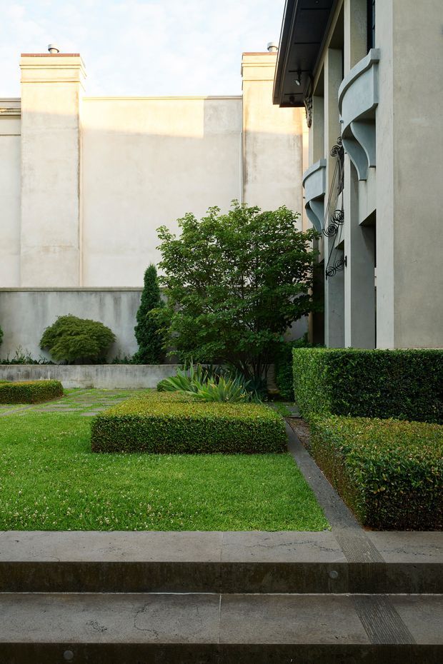 A formal landscaped courtyard viewed from a paved walkway, with clipped rectangular hedges, manicured lawn, ornamental shrubs, and a small tree. The surrounding building has pale rendered walls, tall chimney-like projections, deep eaves, and distinctive curved projecting balcony or façade elements with decorative wrought-iron details. The scene is bright and carefully maintained, with a restrained, symmetrical garden composition.