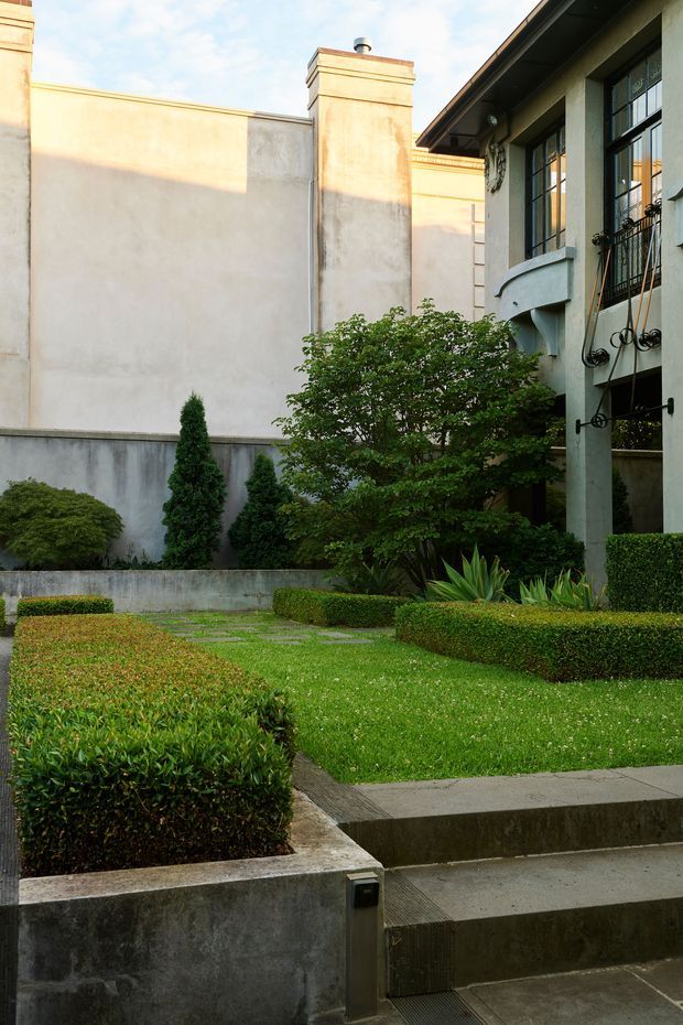 A formal landscaped courtyard beside a multi-storey building, featuring clipped box hedges, manicured lawn, topiary shrubs, stepped concrete paths and raised planting beds. The building has pale rendered walls, dark-framed multi-pane windows, a projecting curved balcony, and decorative wrought-iron balustrades. Warm late-day sunlight creates a refined, enclosed garden atmosphere.