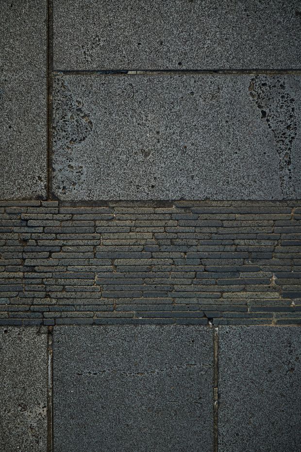Close-up of a dark grey masonry wall composed of large rough-textured rectangular stone or concrete panels, with a horizontal band of narrow dark brickwork through the centre. The surface has visible joints, weathering, and an industrial, heavily textured appearance.