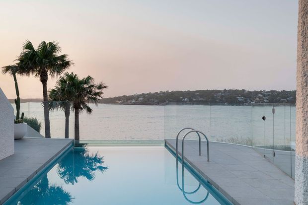 A tranquil waterfront pool terrace overlooking a sheltered bay, with a narrow rectangular swimming pool, stainless-steel ladder, pale stone paving, frameless glass balustrade, and sculptural palm trees. The scene has soft dusk light and a refined coastal-resort character.