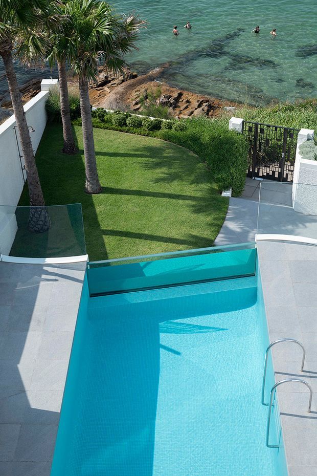 Elevated view of a turquoise swimming pool beside a landscaped lawn and palm trees, with pale paved terraces, white rendered walls, glass pool fencing, and a black metal gate. The property overlooks a rocky waterfront with clear blue-green water and swimmers.