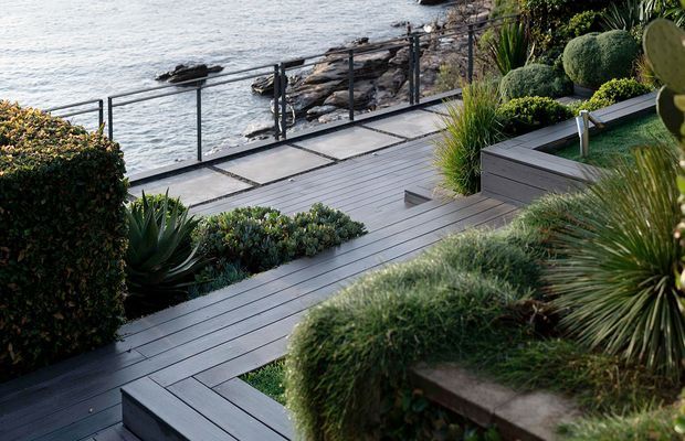 A landscaped coastal terrace overlooking rocky shoreline and the sea. Dark grey timber-look decking forms stepped platforms and pathways, bordered by low concrete or stone retaining edges, glass and metal balustrades, clipped hedges, succulents, grasses and sculptural coastal planting.