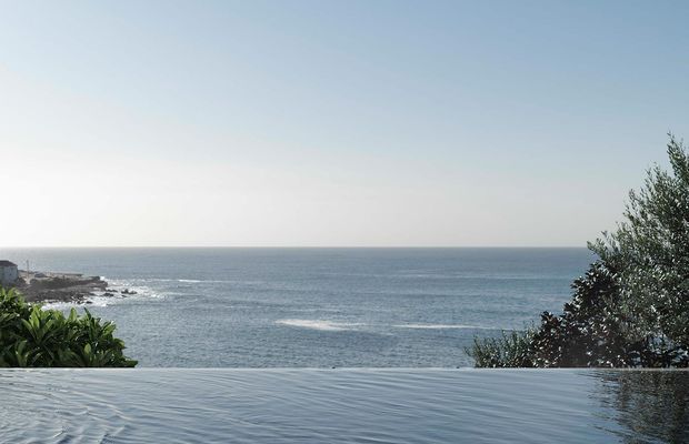 A tranquil ocean vista viewed from the edge of an infinity pool or reflective water feature. The image shows a calm blue sea, rocky shoreline, coastal vegetation, and a pale sky, with the pool surface visually blending into the horizon.