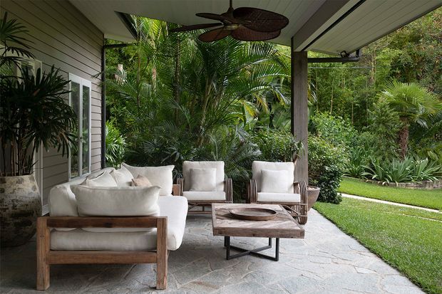 A covered outdoor lounge terrace furnished with white-cushioned timber armchairs and sofas around a rustic wood coffee table. The space has a slatted ceiling with a ceiling fan, grey horizontal wall cladding, large glazed doors, natural stone paving, and dense tropical garden planting beyond.