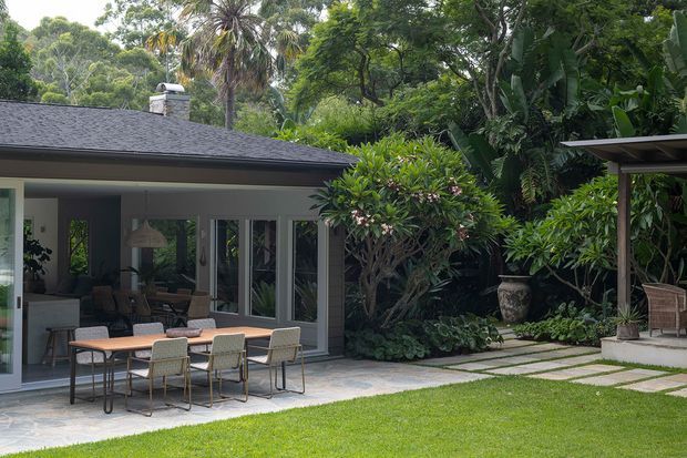 A lush tropical courtyard garden adjoining a single-storey contemporary home. The covered outdoor dining terrace has a timber dining table and woven chairs, with large glazed doors opening to the interior. Stepping stones cross the lawn toward a roofed outdoor structure, surrounded by dense palms and tropical planting.