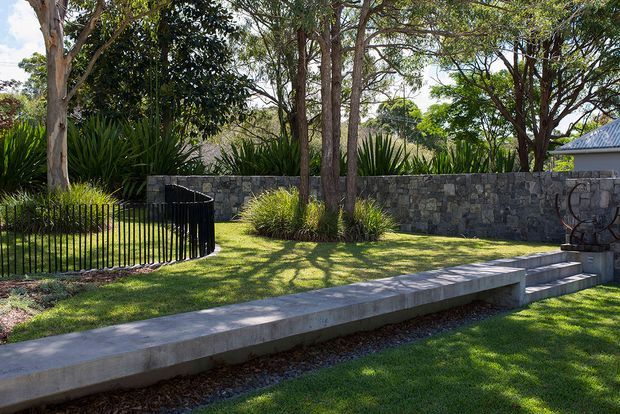 A landscaped outdoor garden area with mature trees, clipped lawn, ornamental grasses and agave-like planting, enclosed by a dark stone masonry wall. A long concrete bench and stepped platform run across the foreground, with black metal railings and a small corrugated metal roof visible at the edge of the garden.