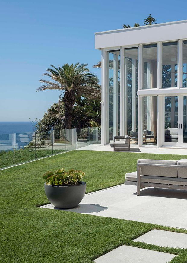 A contemporary coastal home exterior with a crisp white flat-roofed structure and extensive floor-to-ceiling glazing. The image shows a manicured lawn, pale concrete stepping slabs, glass balustrading, outdoor lounge furniture, and a large planter. Palm trees and ocean views are visible beyond the property, creating a bright, open seaside setting.