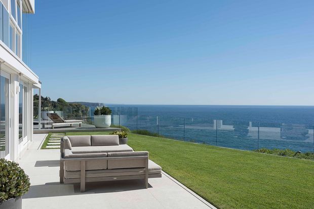 A modern coastal residence viewed from an expansive outdoor terrace overlooking the ocean. The scene features white rendered walls, extensive glazing, clear glass balustrades, pale paving, a neatly landscaped lawn, and a neutral outdoor lounge setting.